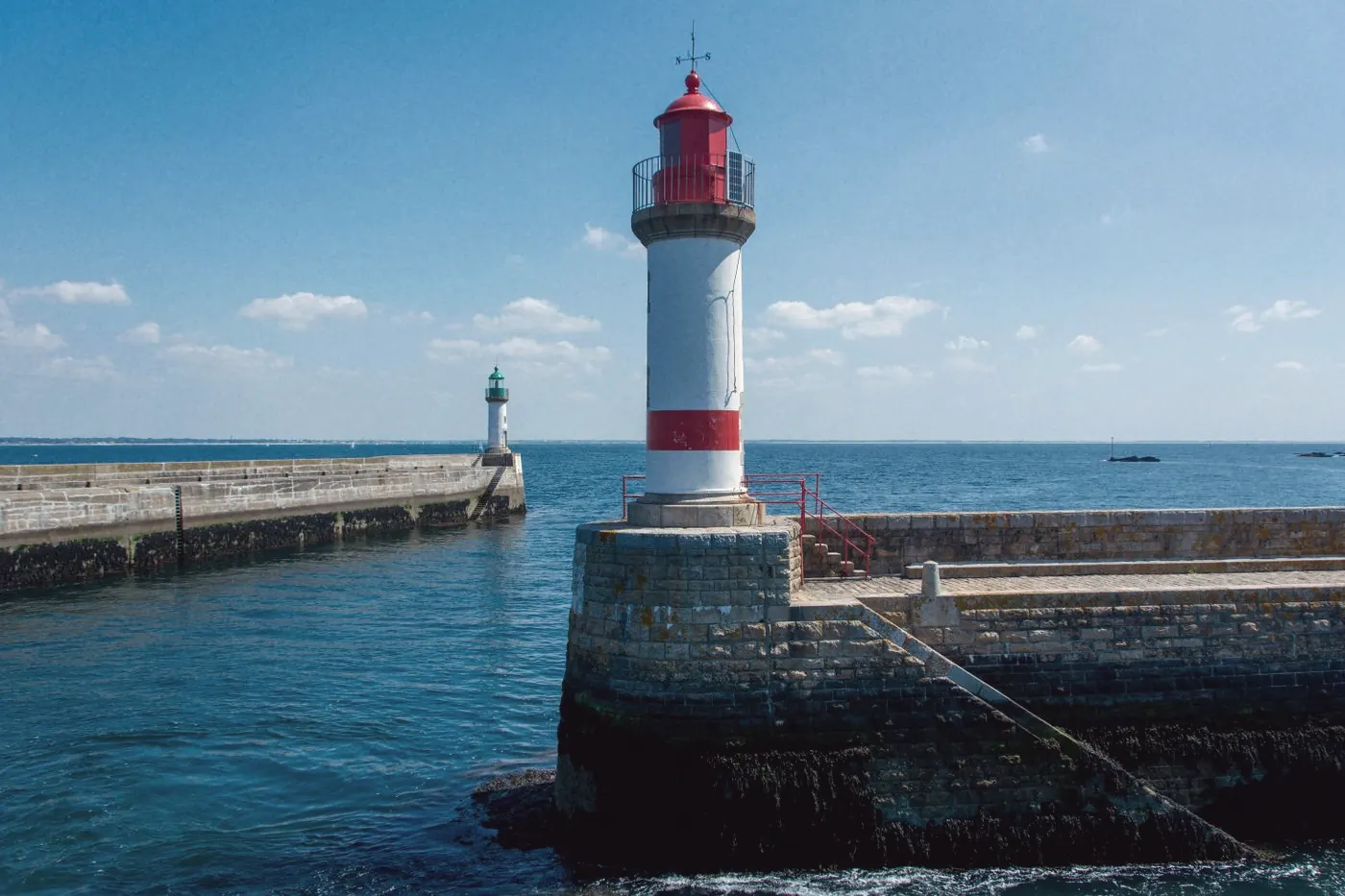 A red and white lighthouse on a stone pier beside a calm blue sea