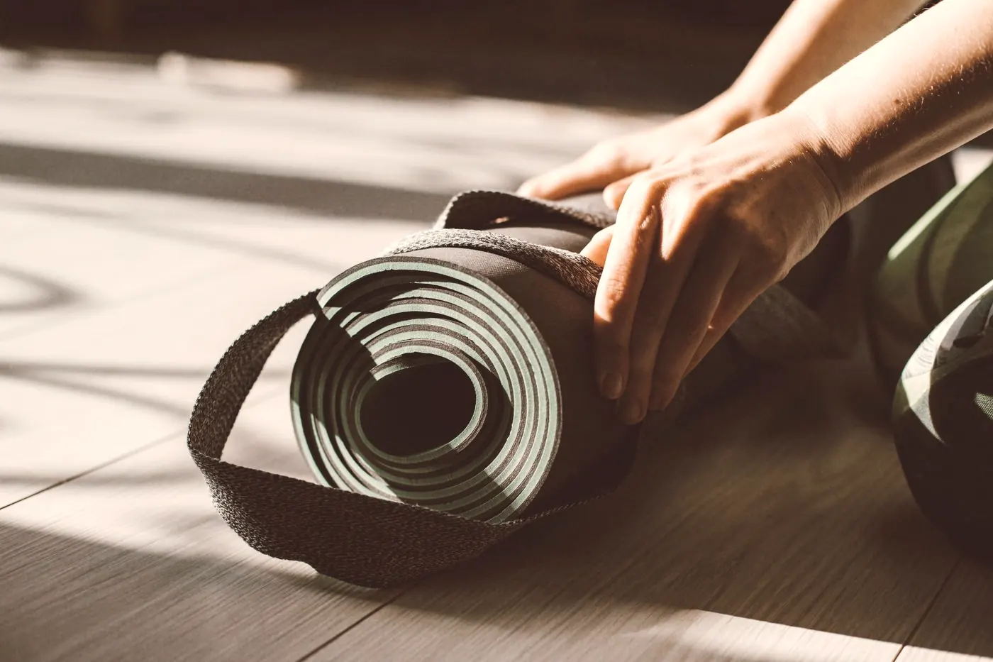 Hands rolling up a yoga mat on a sunlit wooden floor, part of a daily routine