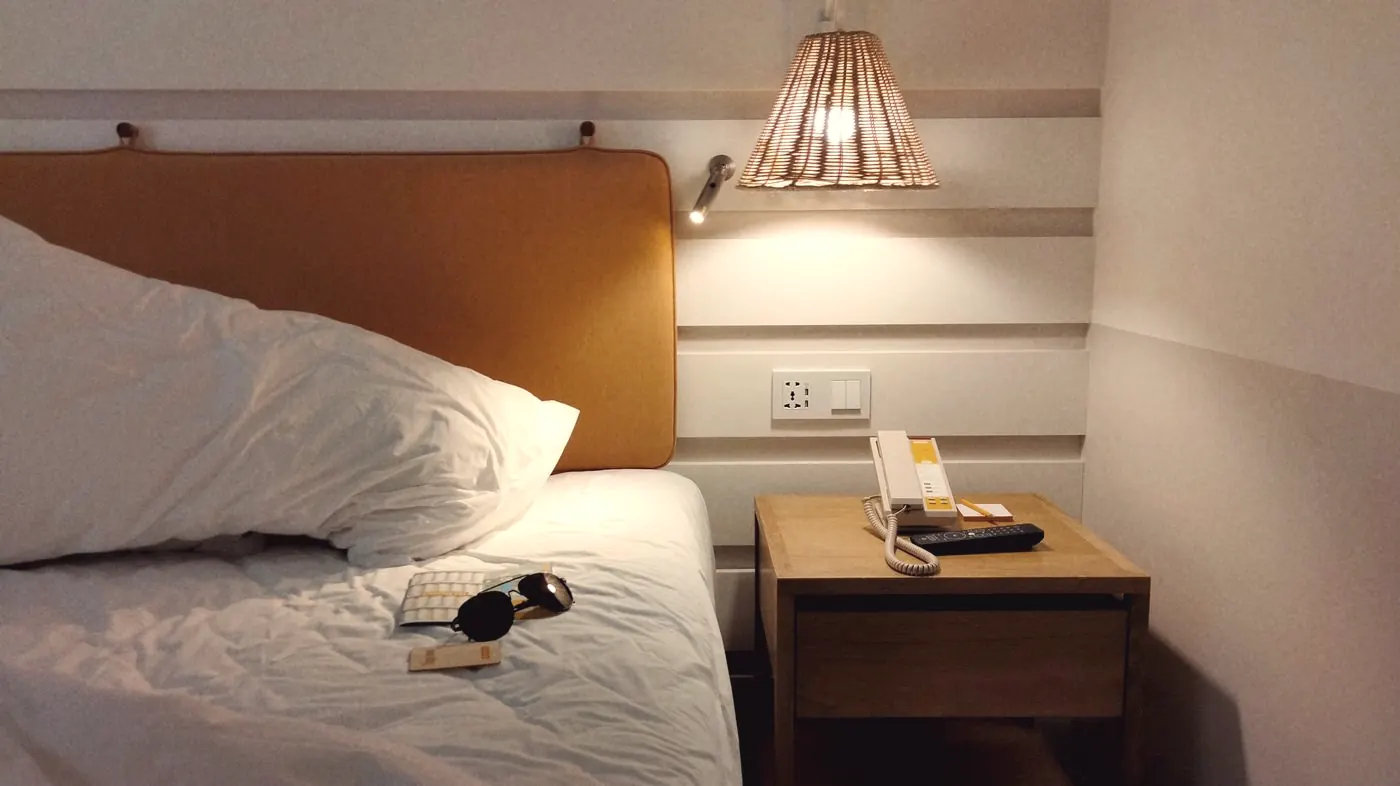 A lamp-lit bedside table with a phone next to a hotel bed late in the evening