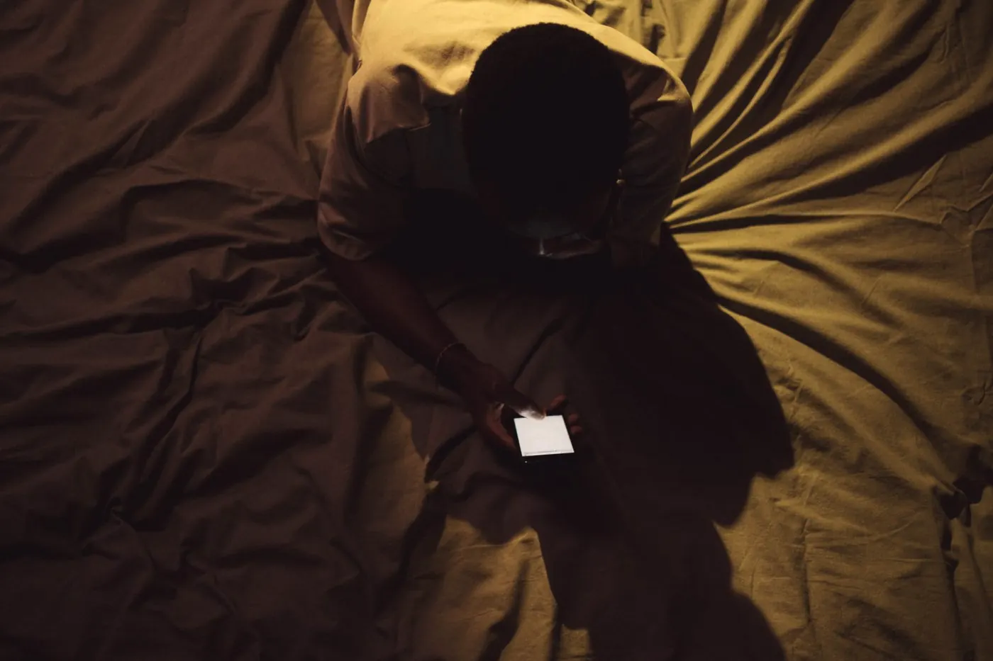 An overhead view of someone in shadow checking a phone on a bed late at night