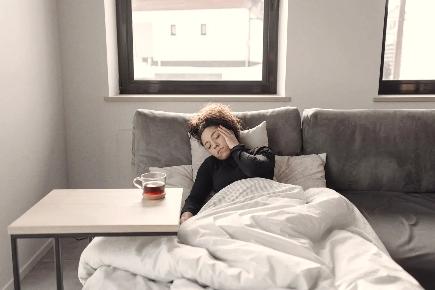 A person resting on a sofa under a blanket with a cup of tea while feeling unwell