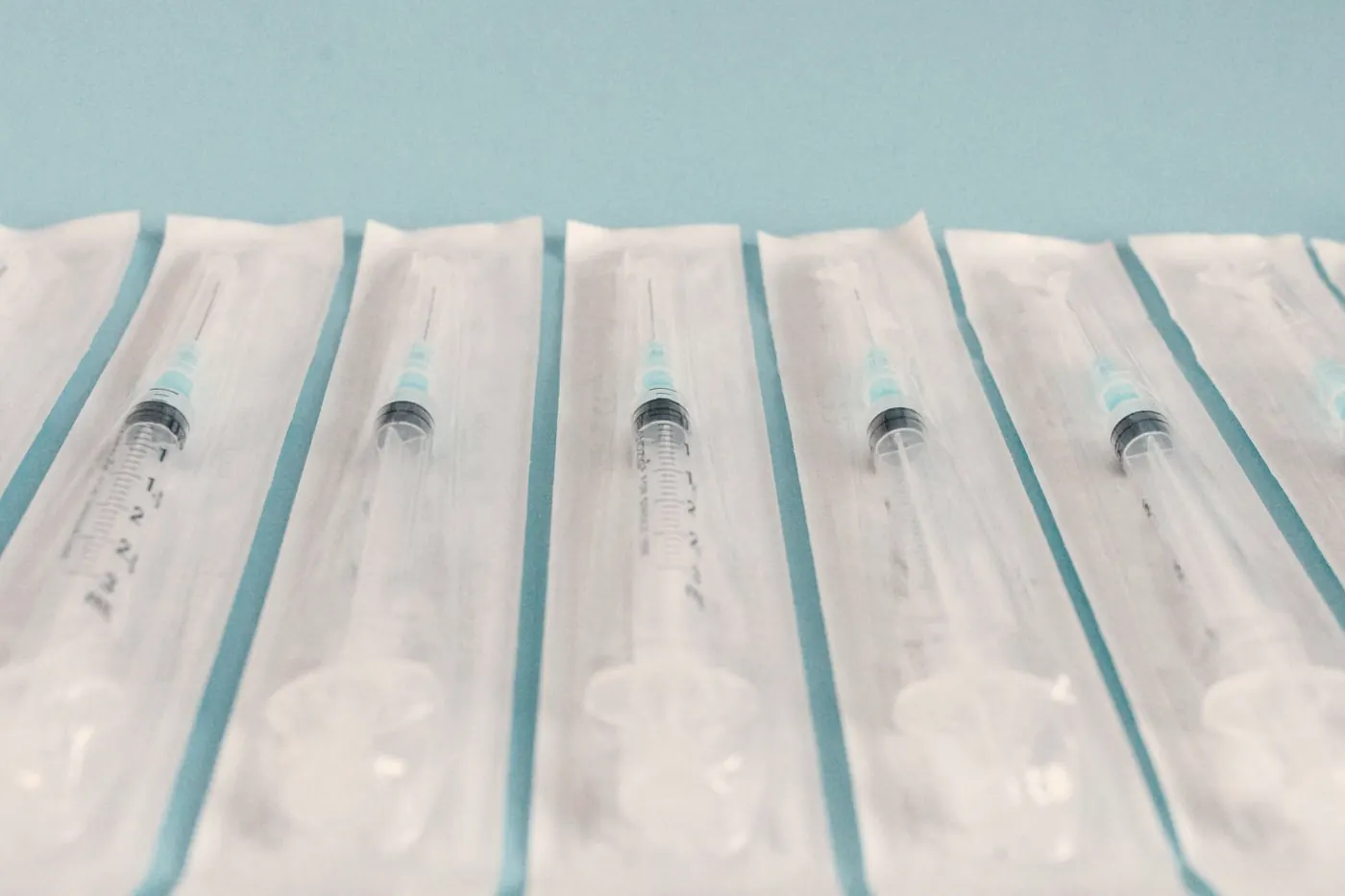 Unused syringes still sealed in their sterile packaging, lined up on a light blue surface