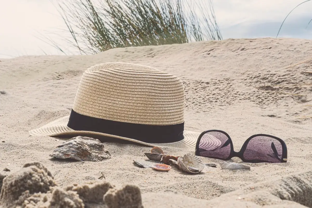 A straw hat and sunglasses resting on a sandy beach beside a few seashells