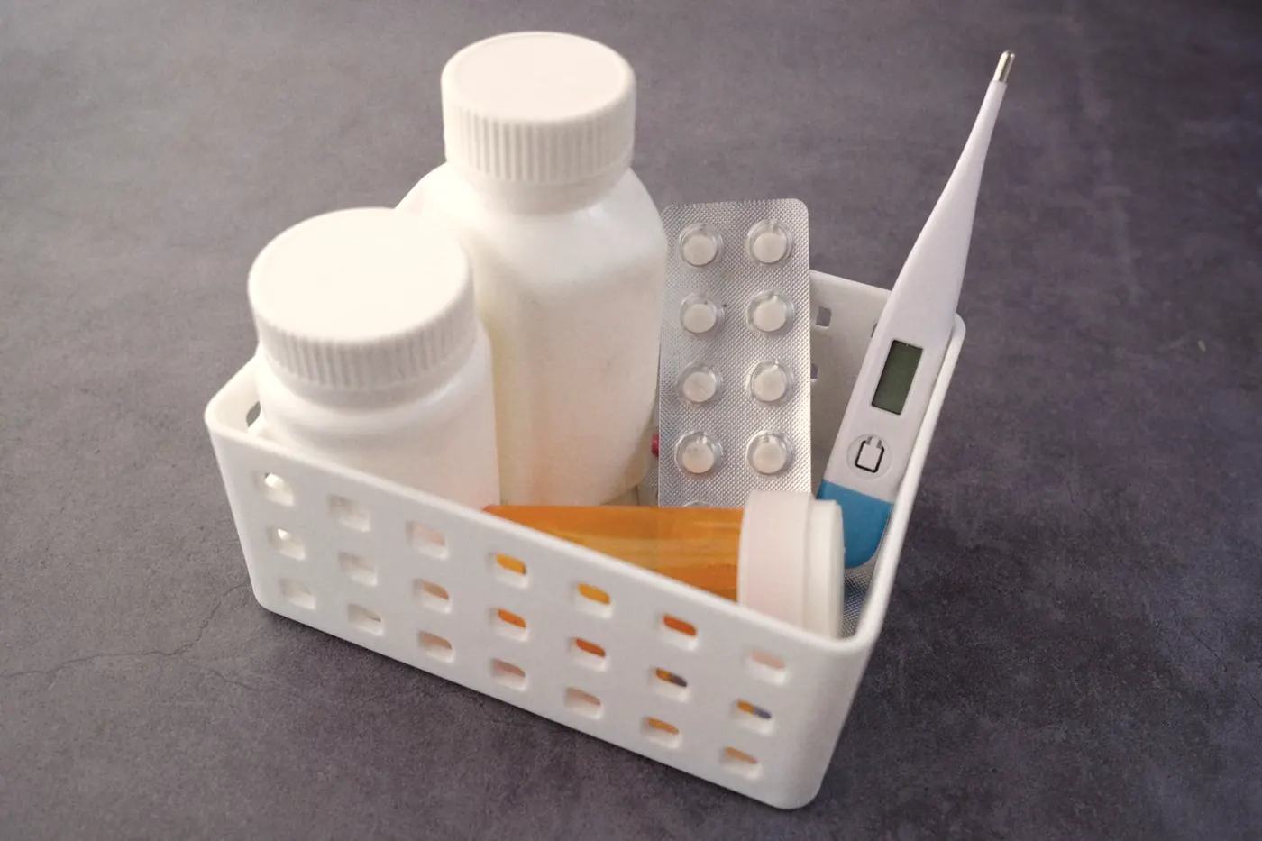 A small basket with a digital thermometer, medicine bottles and tablets on a grey table